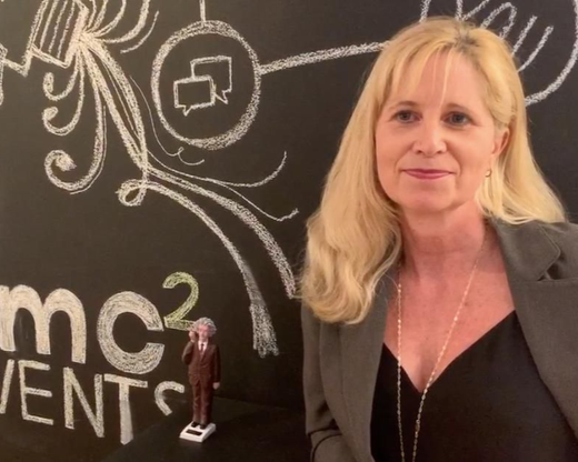 Blonde woman in a blazer standing before a chalkboard-style event backdrop, next to a small trophy