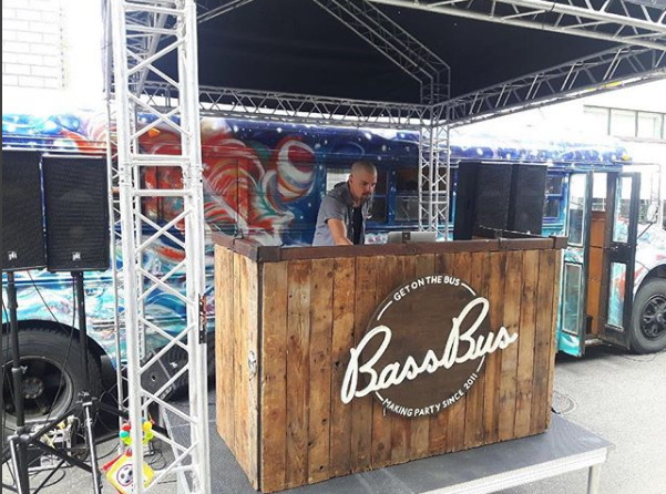 A man is standing behind a wooden bar in front of a bus.