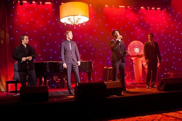 A group of men singing on a stage with a piano in the background