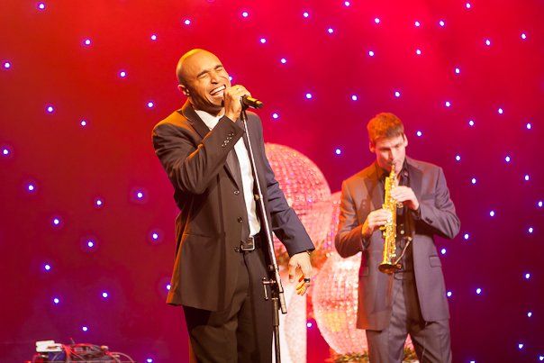 A man is singing into a microphone on a stage while another man plays a saxophone.