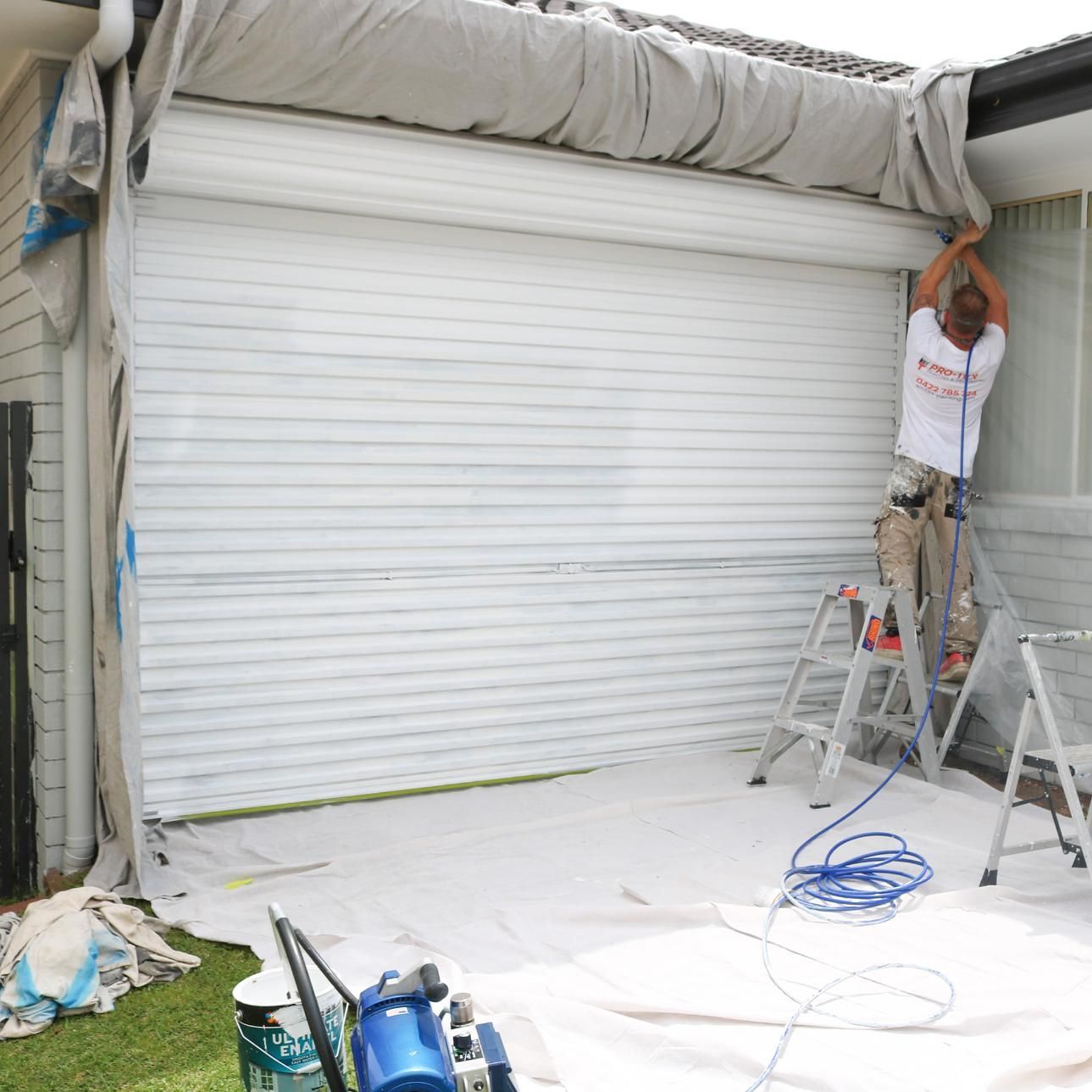 A Man On A Ladder Is Painting A Garage Door — APEX Painting & Maintenance In Primbee, NSW