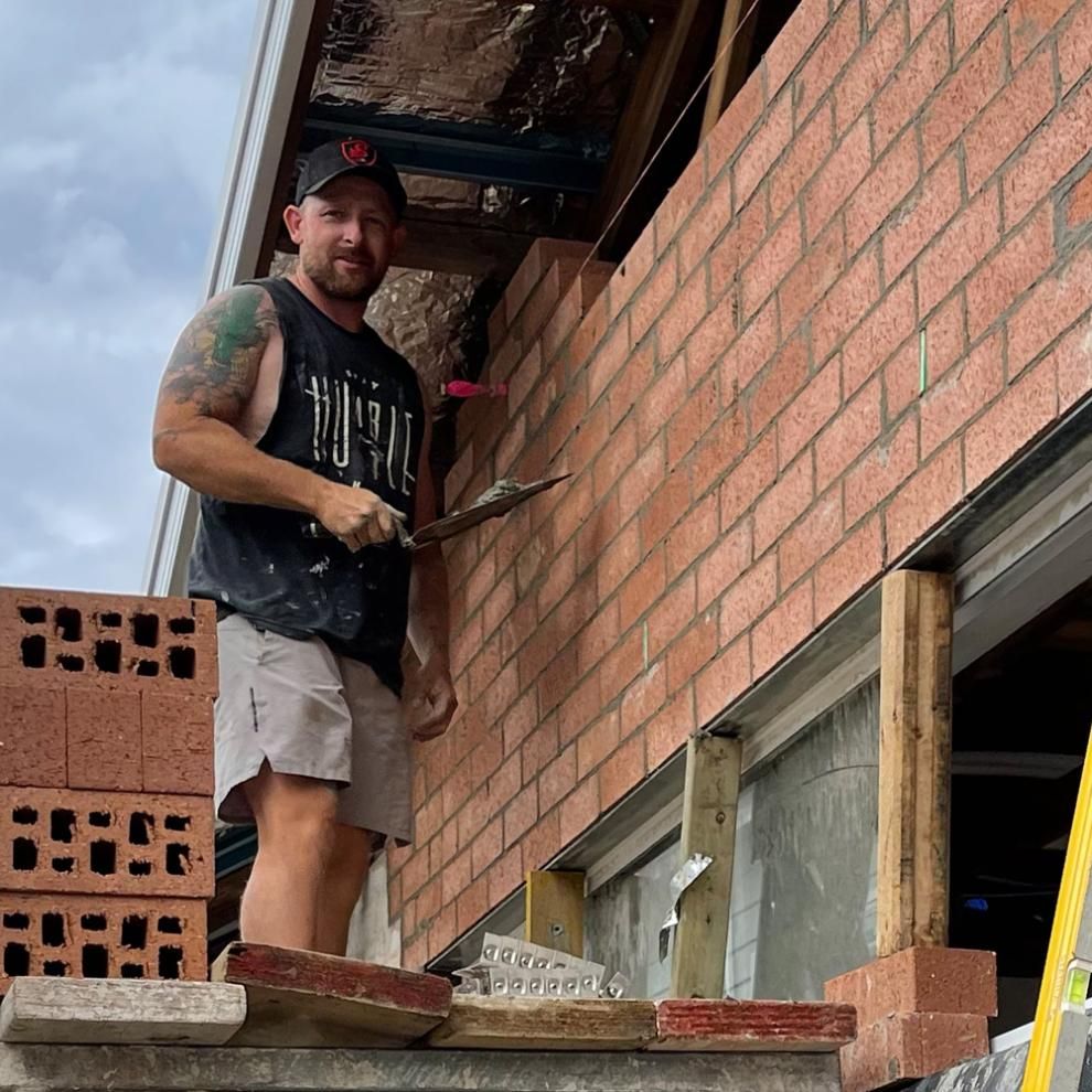 A Man Is Standing On A Brick Wall Holding A Trowel — APEX Painting & Bricklaying In Primbee, NSW