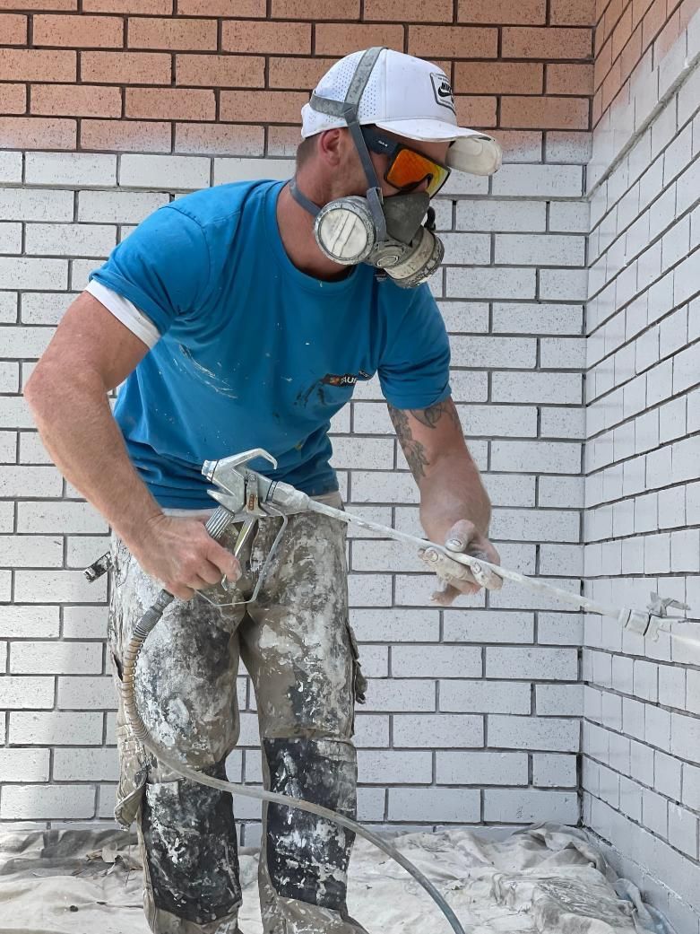 A Man Wearing A Gas Mask Is Spraying Paint On A Brick Wall — APEX Painting & Maintenance In Primbee, NSW