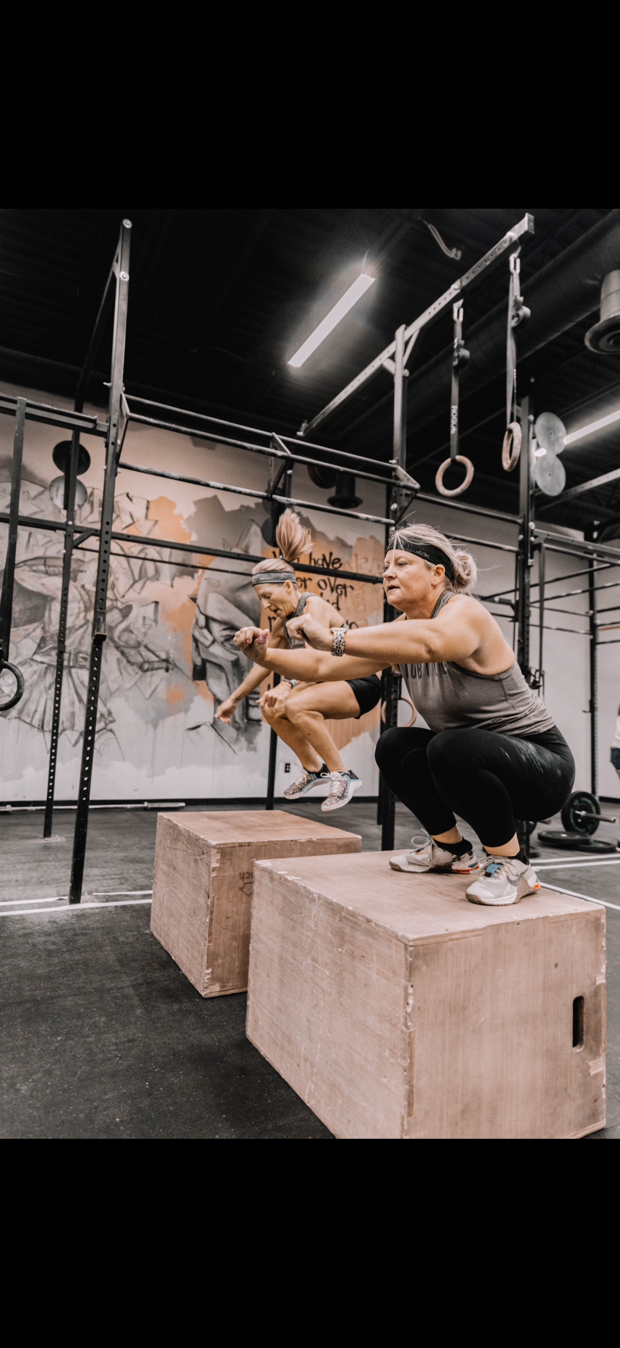 Person squatting on a wooden box in a gym, reaching toward a hanging ring among rigged equipment.