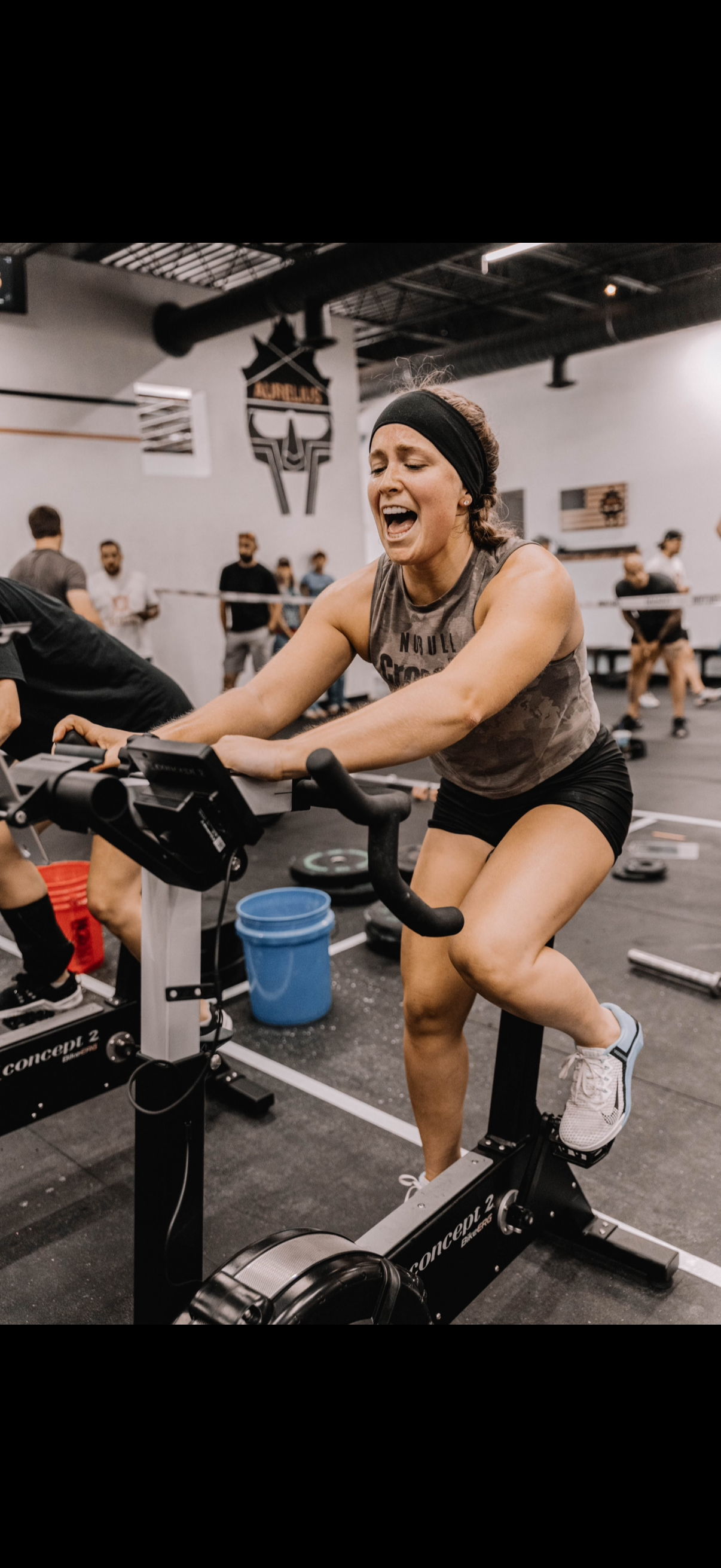 Woman cycling intensely on a stationary bike in a gym, smiling with effort, other exercisers blurred in background