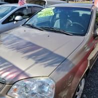 Tan sedan parked outdoors at a car lot, viewed from the front-left with a price sticker on the windshield