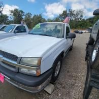 White pickup truck parked in a lot beside other vehicles, with an American flag visible in the background