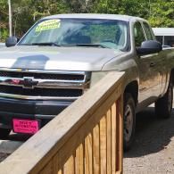 Silver Chevrolet pickup parked by a wooden railing outdoors, with trees in the background.