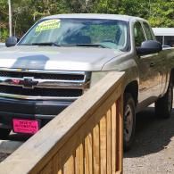 Silver Chevrolet pickup parked beside a wooden railing outdoors in bright sunlight.
