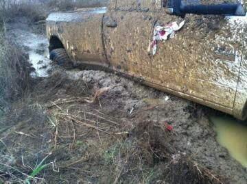 Mud-covered car overturned in a ditch beside muddy water.
