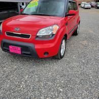 Red Kia Soul parked on gravel, front-left view, with a pink license plate frame.