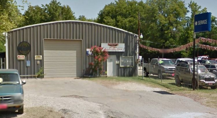 Small industrial warehouse with a roll-up garage door, parked cars, and a blue sign by the driveway.