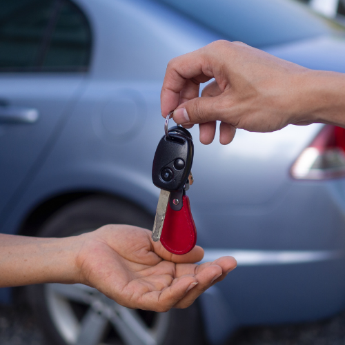 Car and house keys being handed between two people in front of a blue car