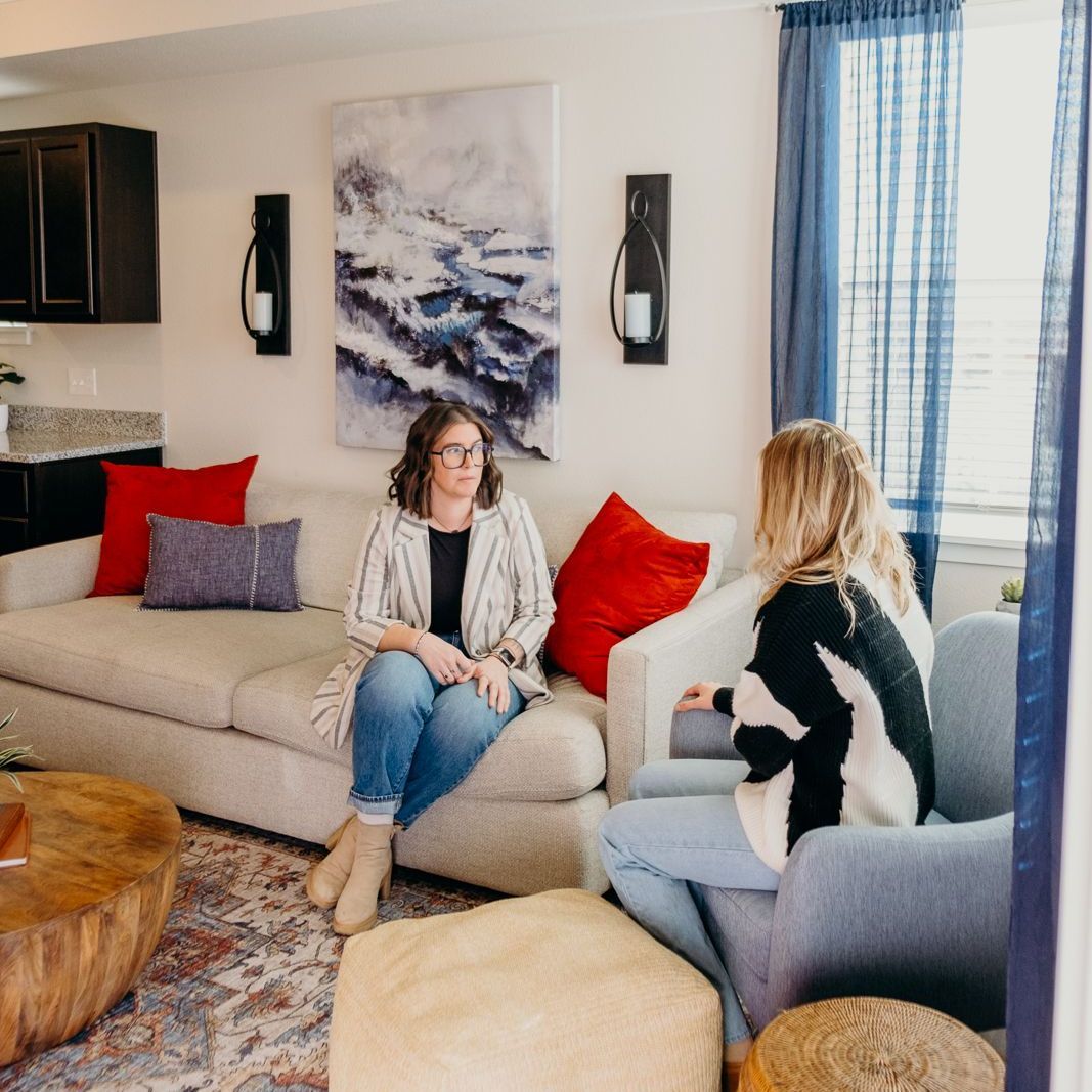 Two women are sitting on a couch in a living room talking to each other.