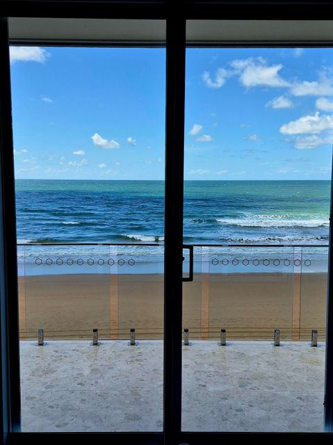 View through a window of a sandy beach, blue ocean, and bright sky with clouds