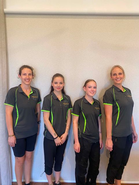 Four people in matching gray and green shirts stand against a plain wall, smiling.