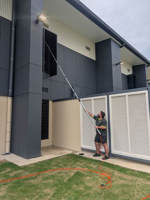 Person washing an upper-story window with a long pole beside a modern house and lawn