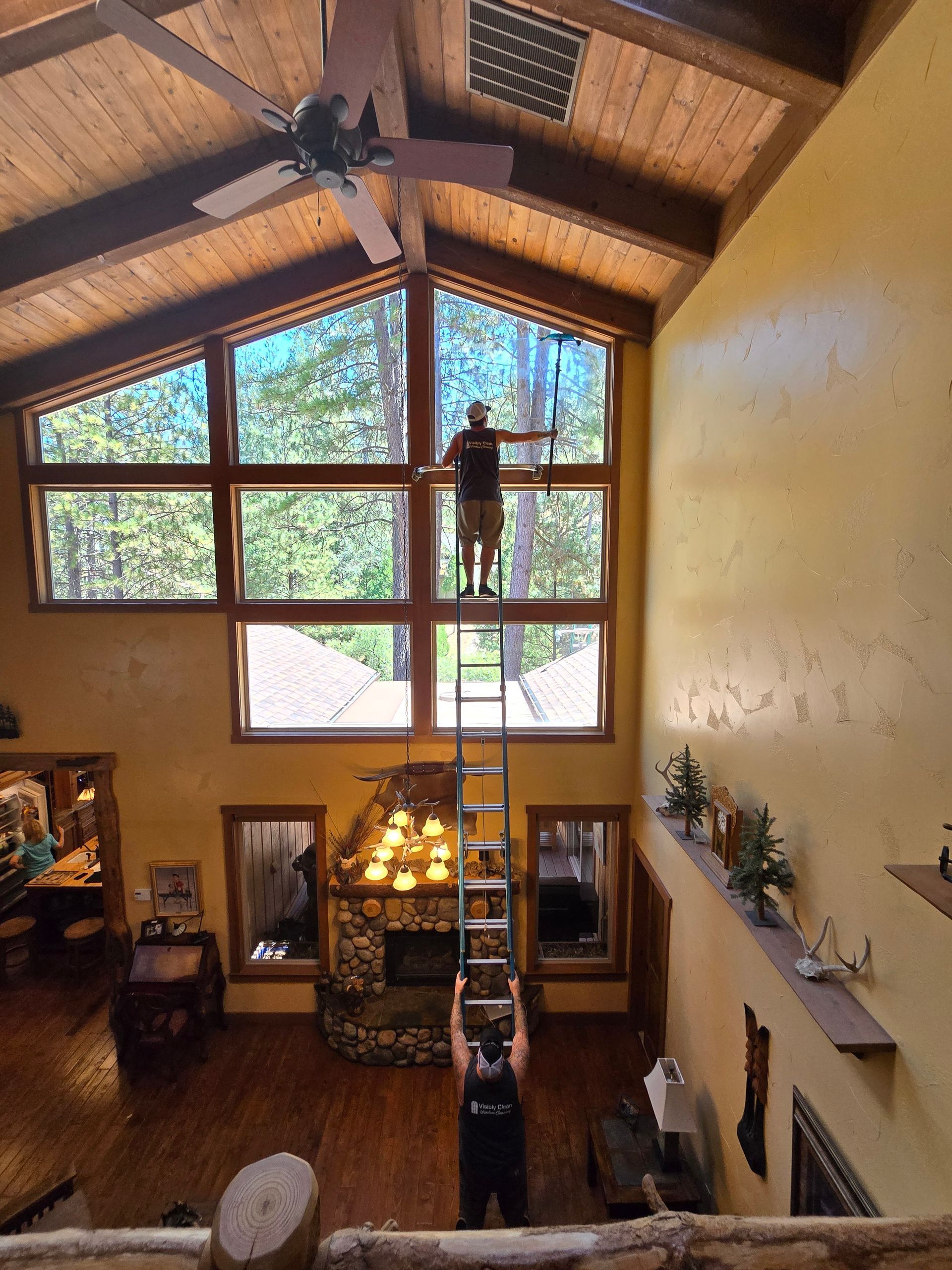 A man is standing on a ladder cleaning a window in a living room.