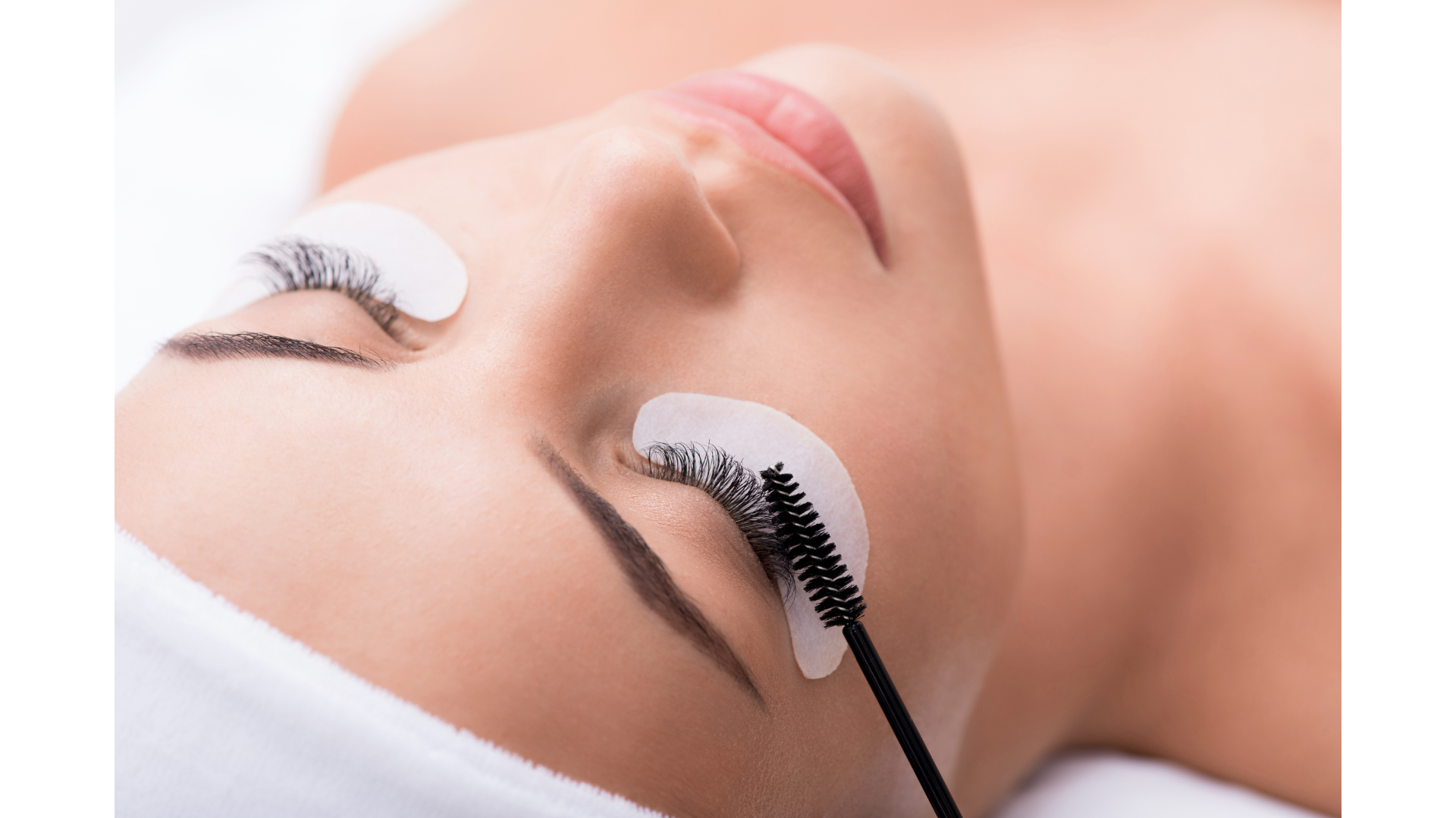 Woman getting eyelash extensions, lying down with under-eye patches. A brush touches the extended lashes.