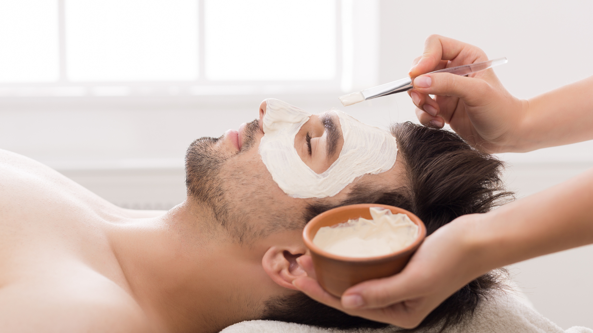 Man receiving facial treatment; esthetician applying mask with brush; spa setting.