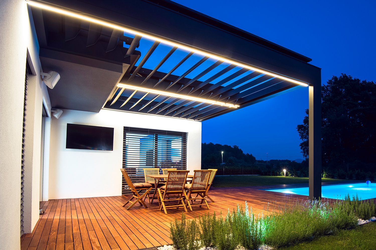 Patio with wooden deck, table, chairs, and pergola with lights, next to a pool at night. Patio with wooden deck, table, chairs, and pergola with lights, next to a pool at night.