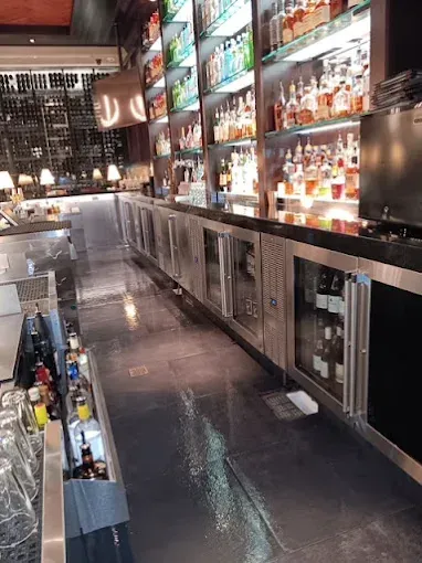 A long, sleek bar with stainless steel coolers, liquor bottles, and glasses, indoors.