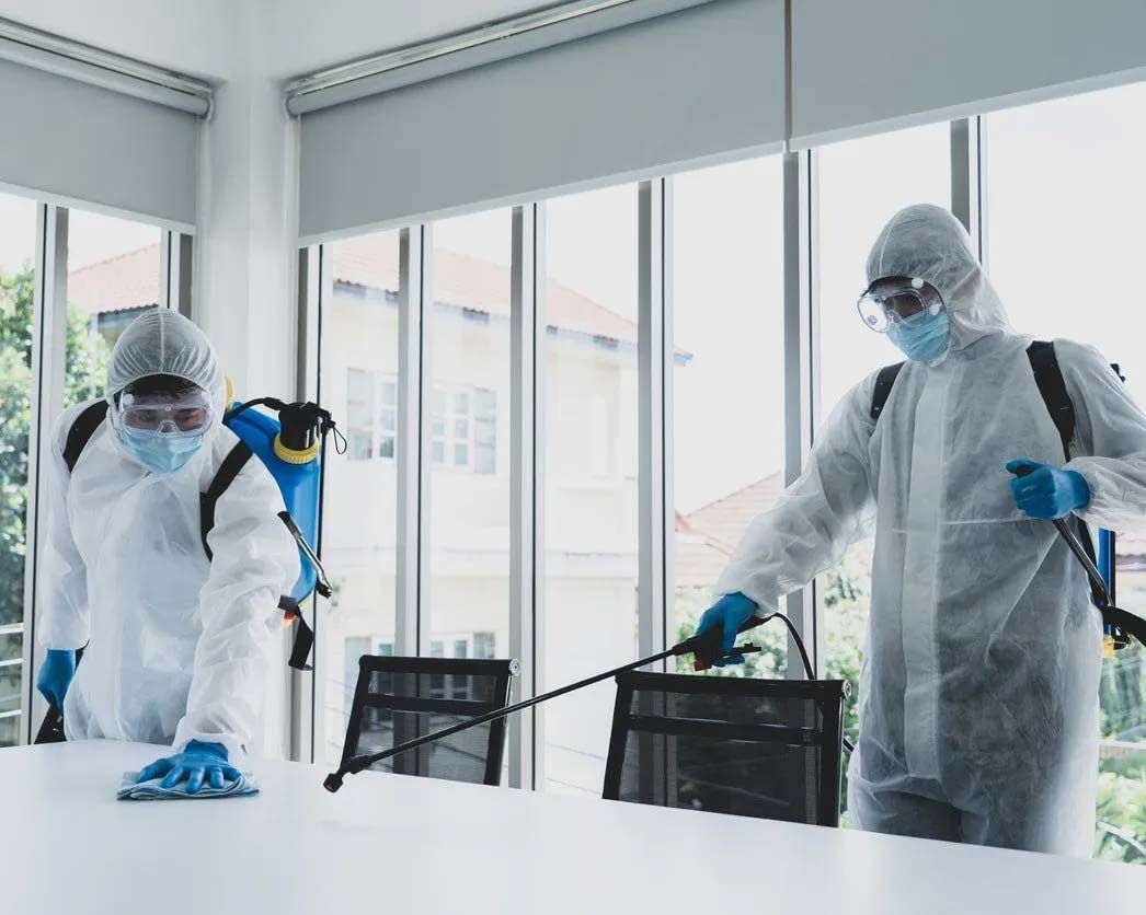 Two people in hazmat suits sanitizing an office with sprayers and cleaning cloths.