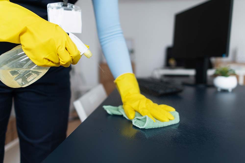 Person cleaning a dark desk with spray bottle and cloth, wearing yellow gloves in office setting.