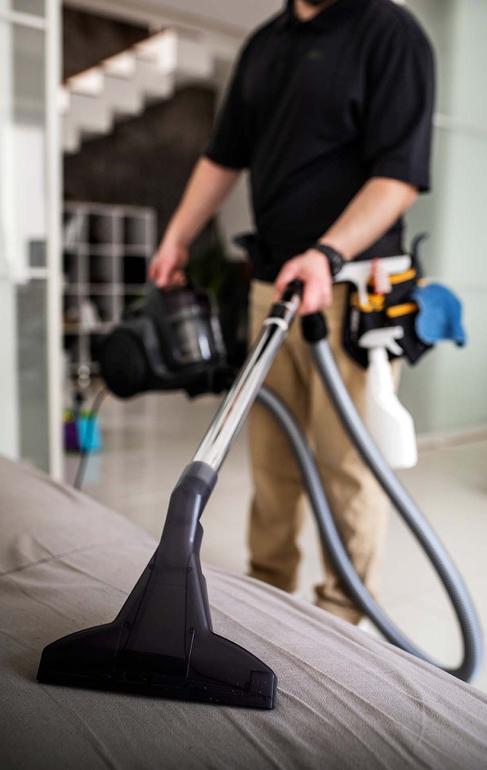 Person vacuuming a sofa, holding a vacuum cleaner and cleaning supplies in an indoor setting.
