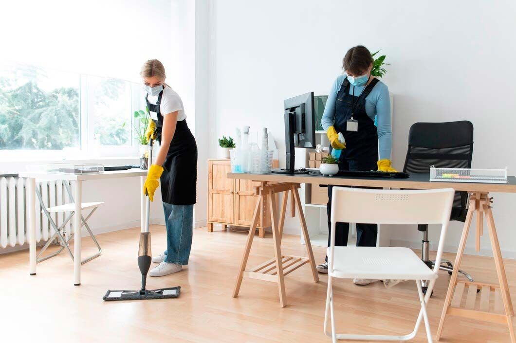 Two people wearing masks and aprons cleaning an office with a mop and spray bottle.