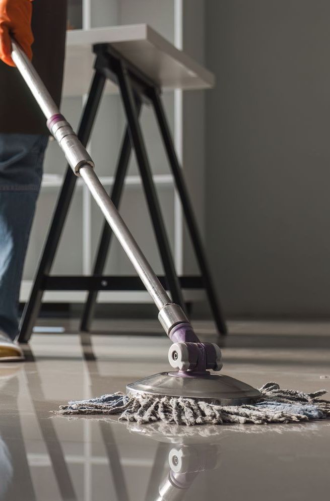Person mopping a glossy floor. They wear gloves, with a shelving unit and wall in background.