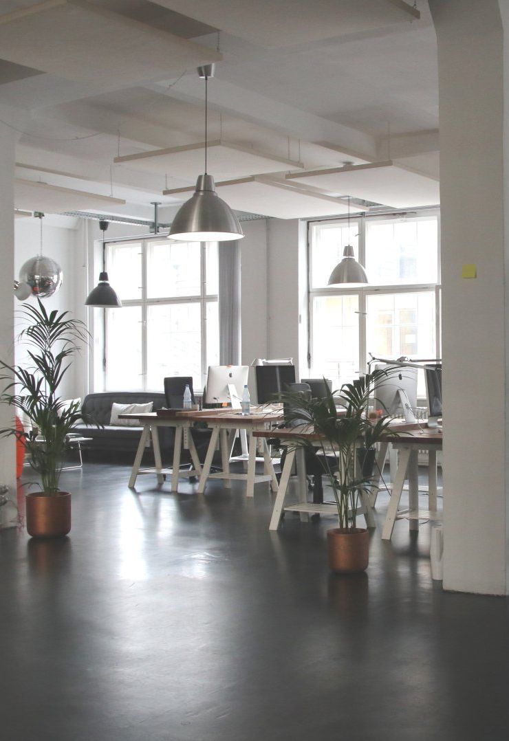 Open-plan office with desks, computers, potted plants, and pendant lights. Windows in the background.