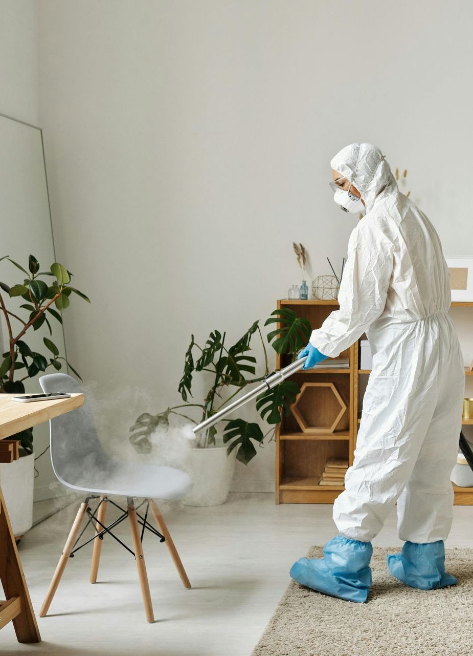 Person in hazmat suit disinfecting a chair with a sprayer indoors.