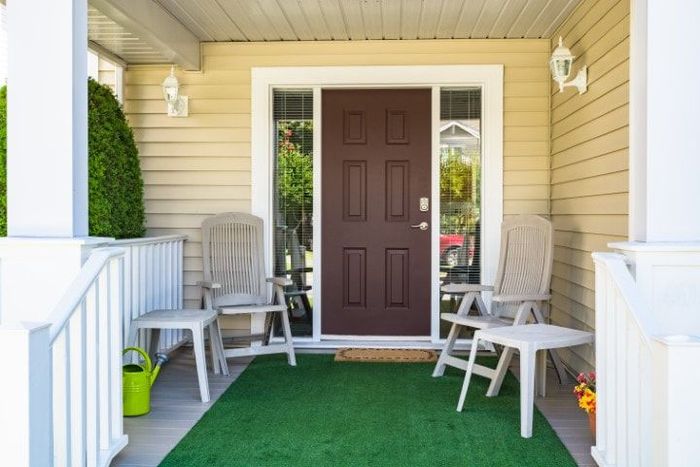 Tan porch with brown door, green rug, white chairs.