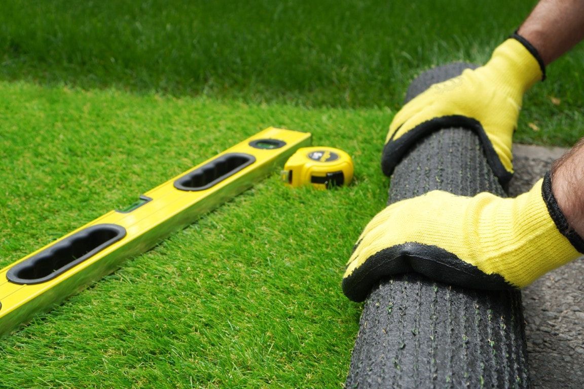Hands in yellow gloves rolling black edging along a lawn, with level and measuring tape nearby.
