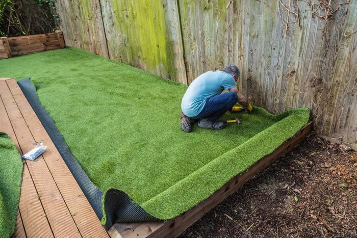 Man installing artificial turf on wooden platform next to a weathered fence.