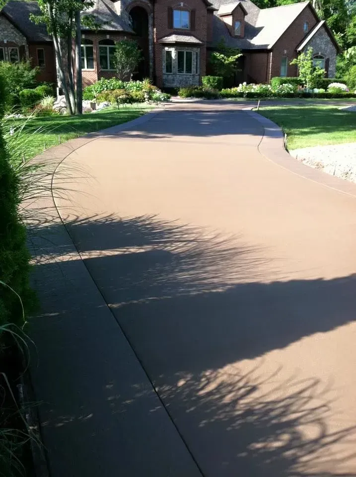 Tan concrete driveway curves toward a large house with bay windows. Sunny day.