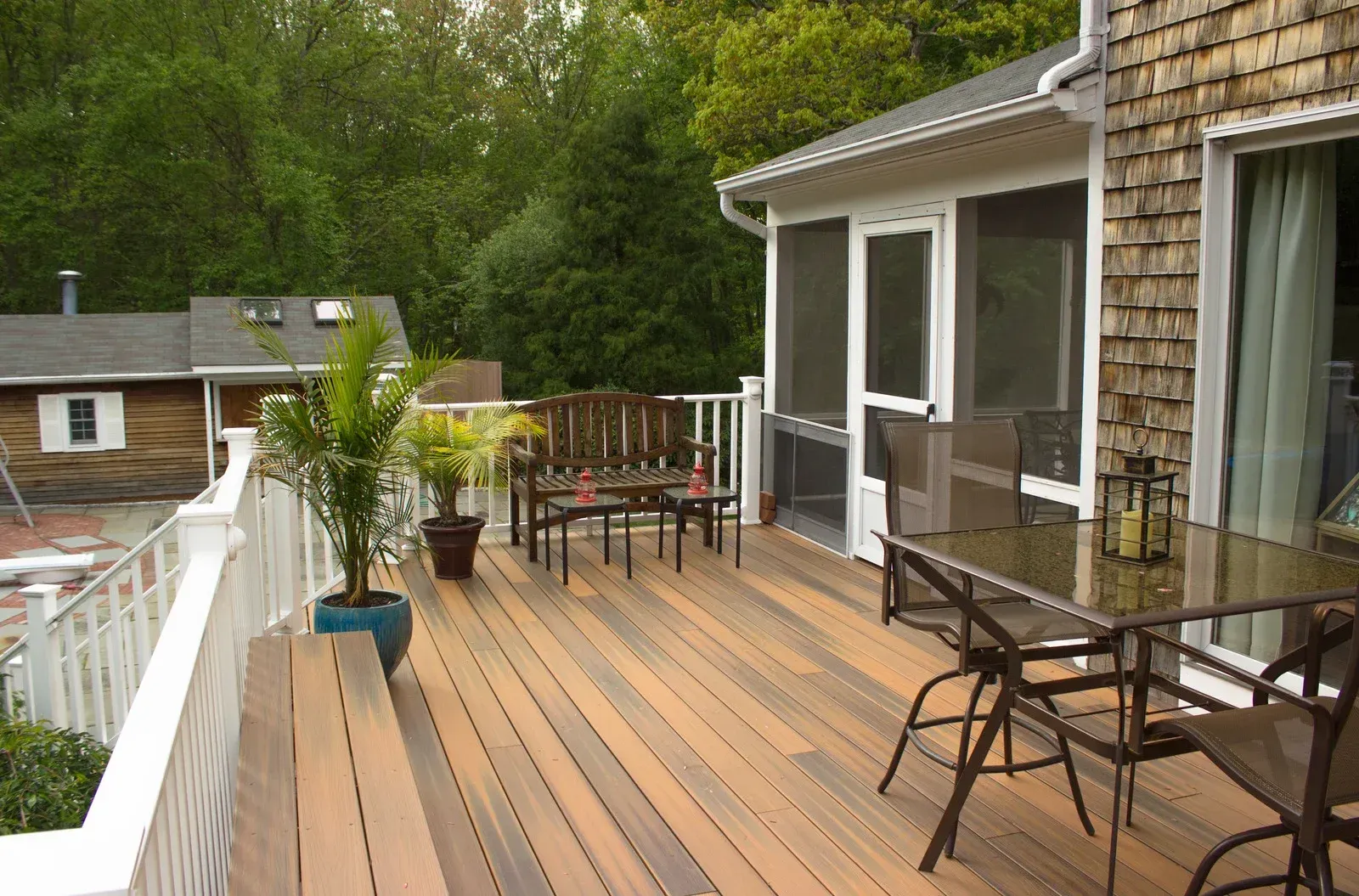 A wooden deck with furniture, potted plants, and a screened porch overlooks a wooded area.