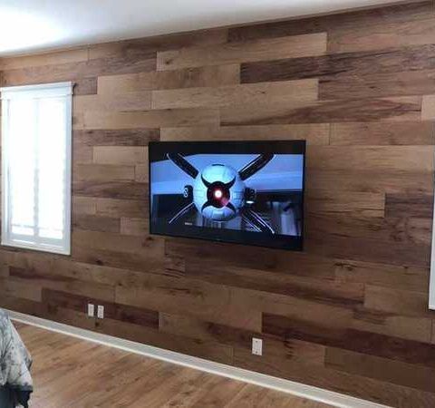 Wall-mounted TV on a wood-paneled living room wall with a window to the left and hardwood floor below