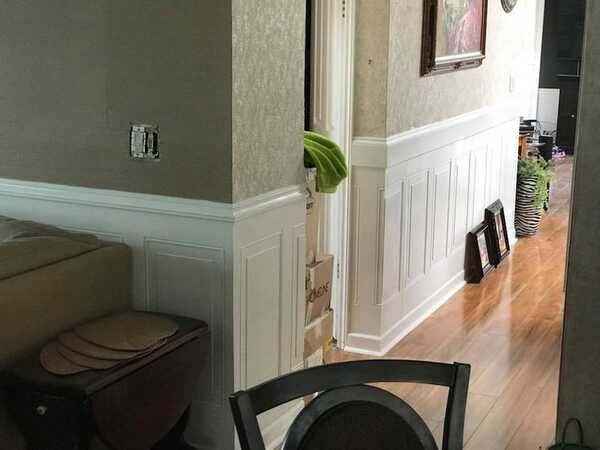 Hallway with white wainscoting, hardwood floor, chairs, and a doorway to a carpeted room with a green cushion