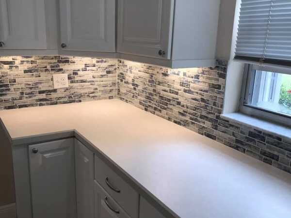 Modern kitchen corner with white countertops, gray cabinets, mosaic backsplash, and a window with blinds.