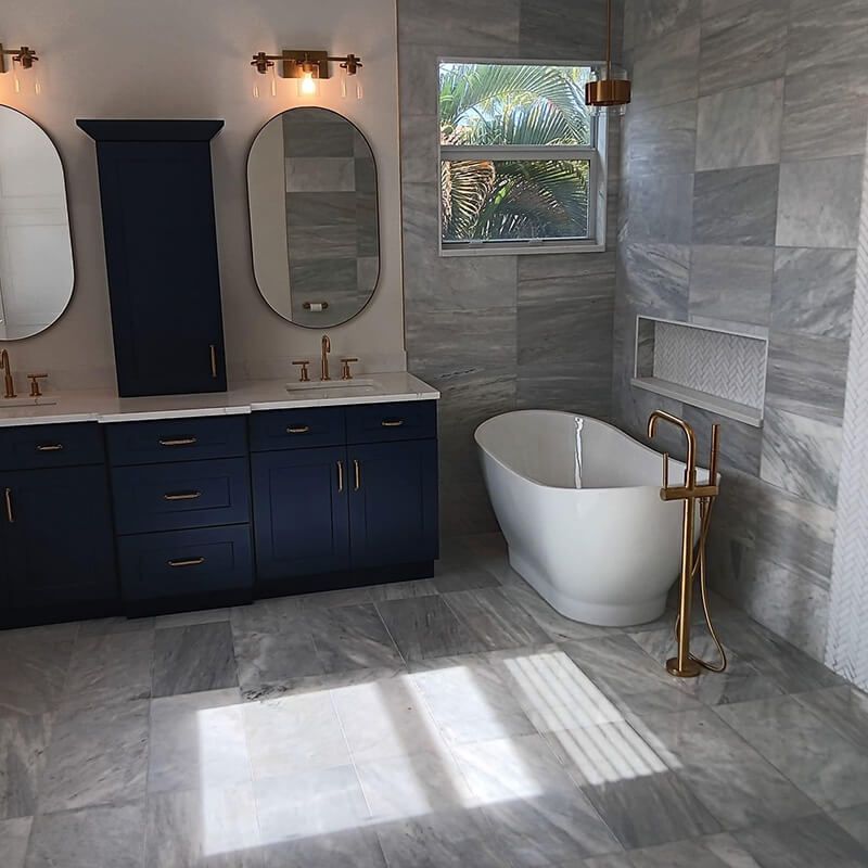 Modern bathroom with navy vanity, oval mirrors, gray tile, and a freestanding white tub by a window