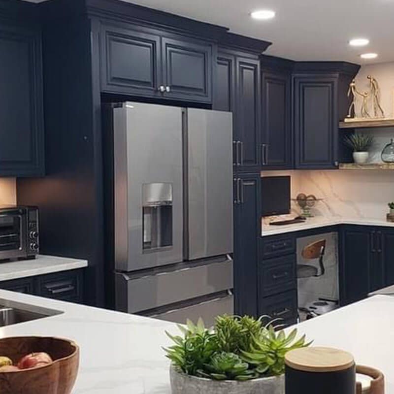 Modern kitchen with dark navy cabinets, stainless steel refrigerator, white countertops, and a small plant in front.