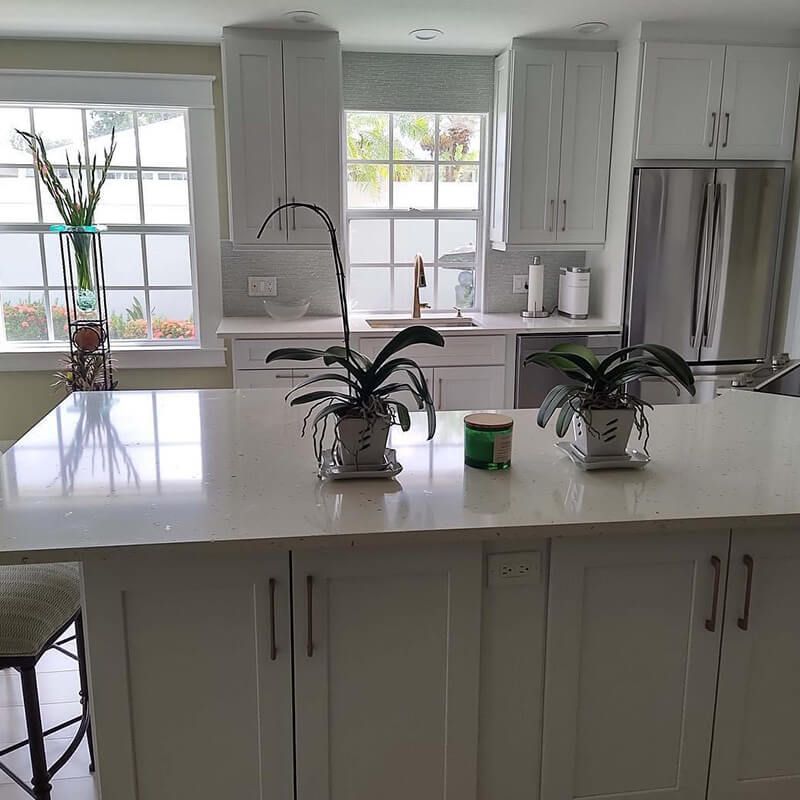 Bright white kitchen with island, potted plants, stainless fridge, and large windows letting in daylight