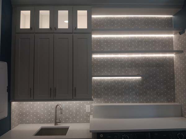 Modern kitchen wall with dark cabinets, sink, and lit open shelves on a patterned backsplash.