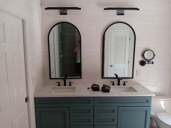 Double vanity bathroom with teal cabinets, two arched mirrors, black sconces, and a white countertop.