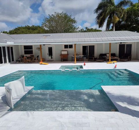 Modern house with turquoise pool, white lounge chair, and palm trees under a blue sky
