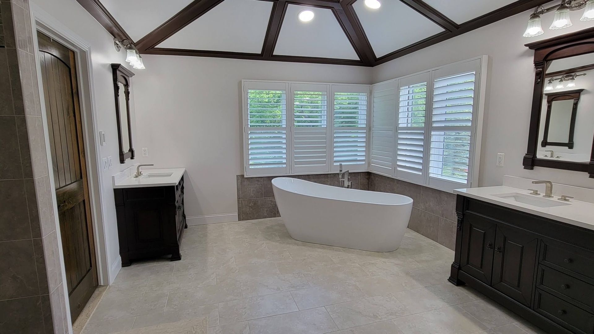 Spacious bathroom with freestanding tub, large windows, exposed beams, and dark wood vanity.