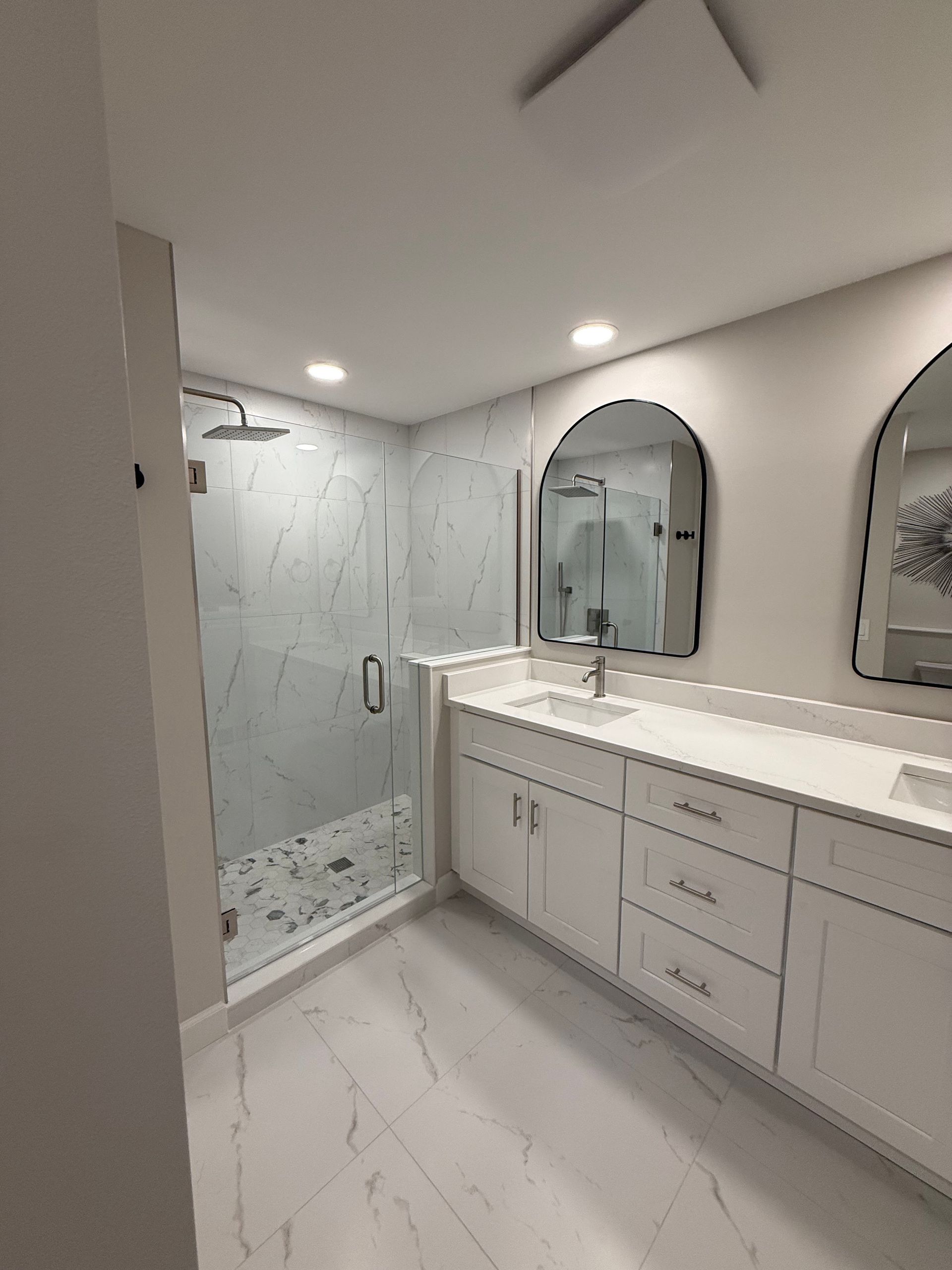 Modern white bathroom with double vanity, large mirrors, glass shower, and marble-style tile.