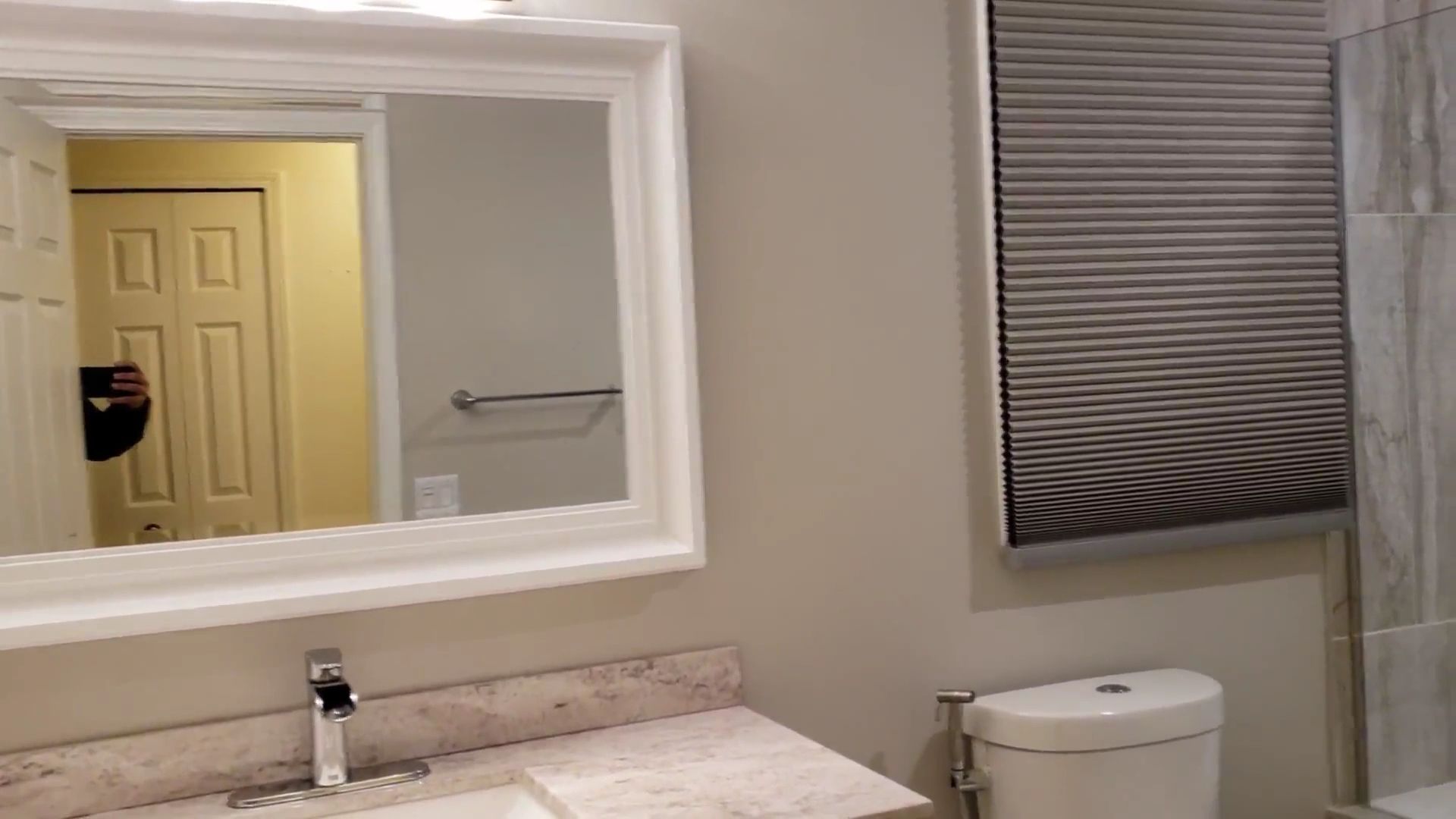 Bathroom vanity with white sink, mirror, toilet, and window blinds on a beige wall.